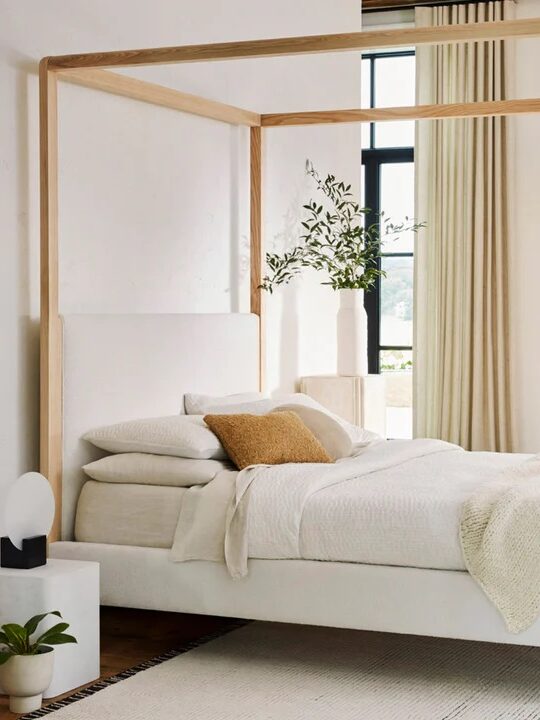 Minimalist bedroom with a wooden canopy bed, white bedding, and a small green ottoman. Beige curtains cover large windows. A potted plant and abstract art add decor.