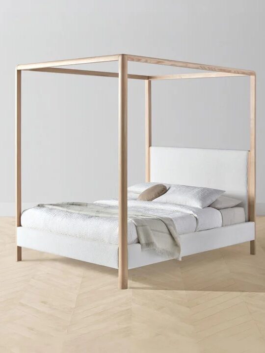 A minimalist canopy bed with a wooden frame, white bedding, and a single pillow in a bright room with light wood flooring.