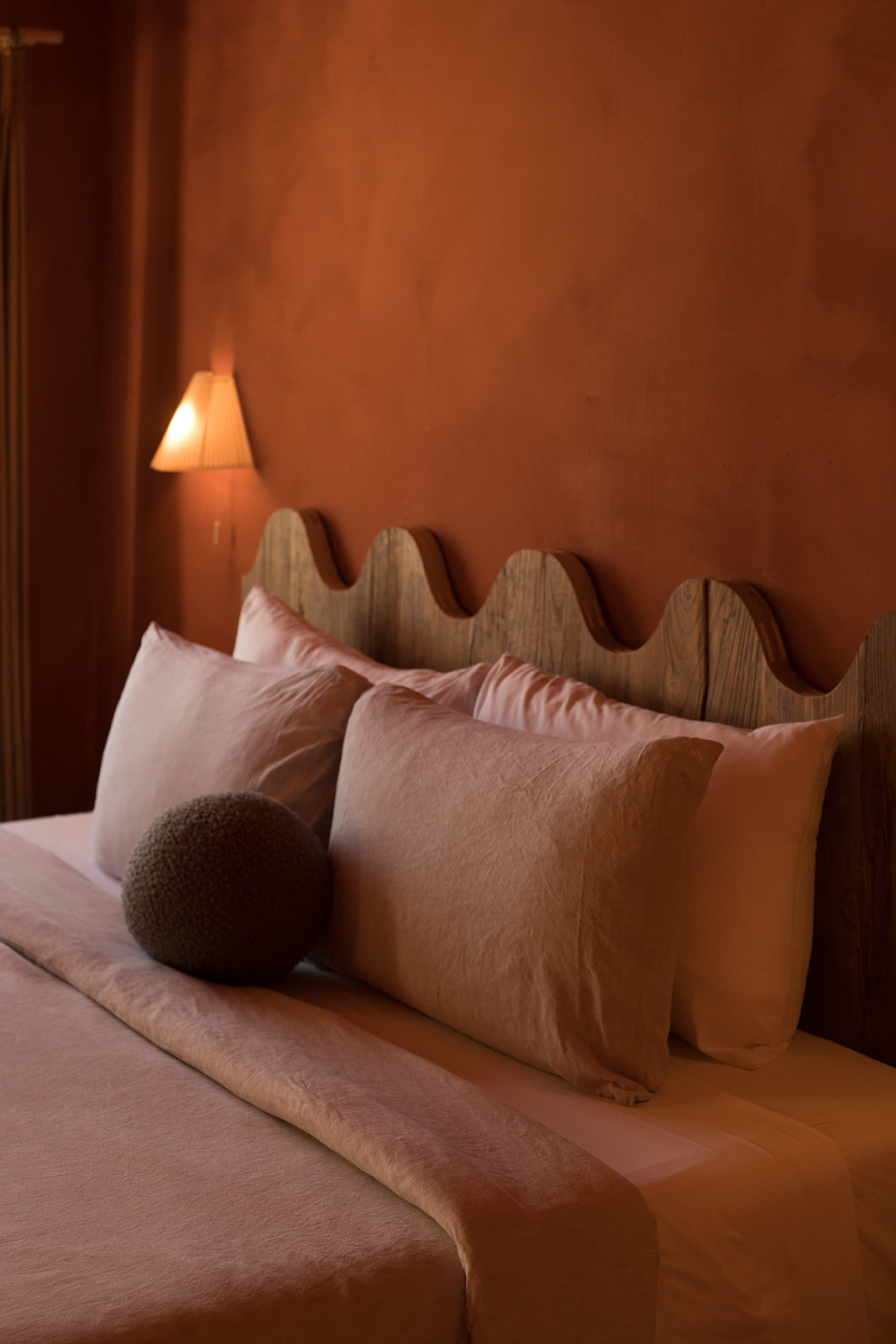 A cozy, warmly-lit bedroom with a uniquely designed wooden headboard, two large pillows, a round cushion, and a small wall lamp. The walls are painted in a rich terracotta color.