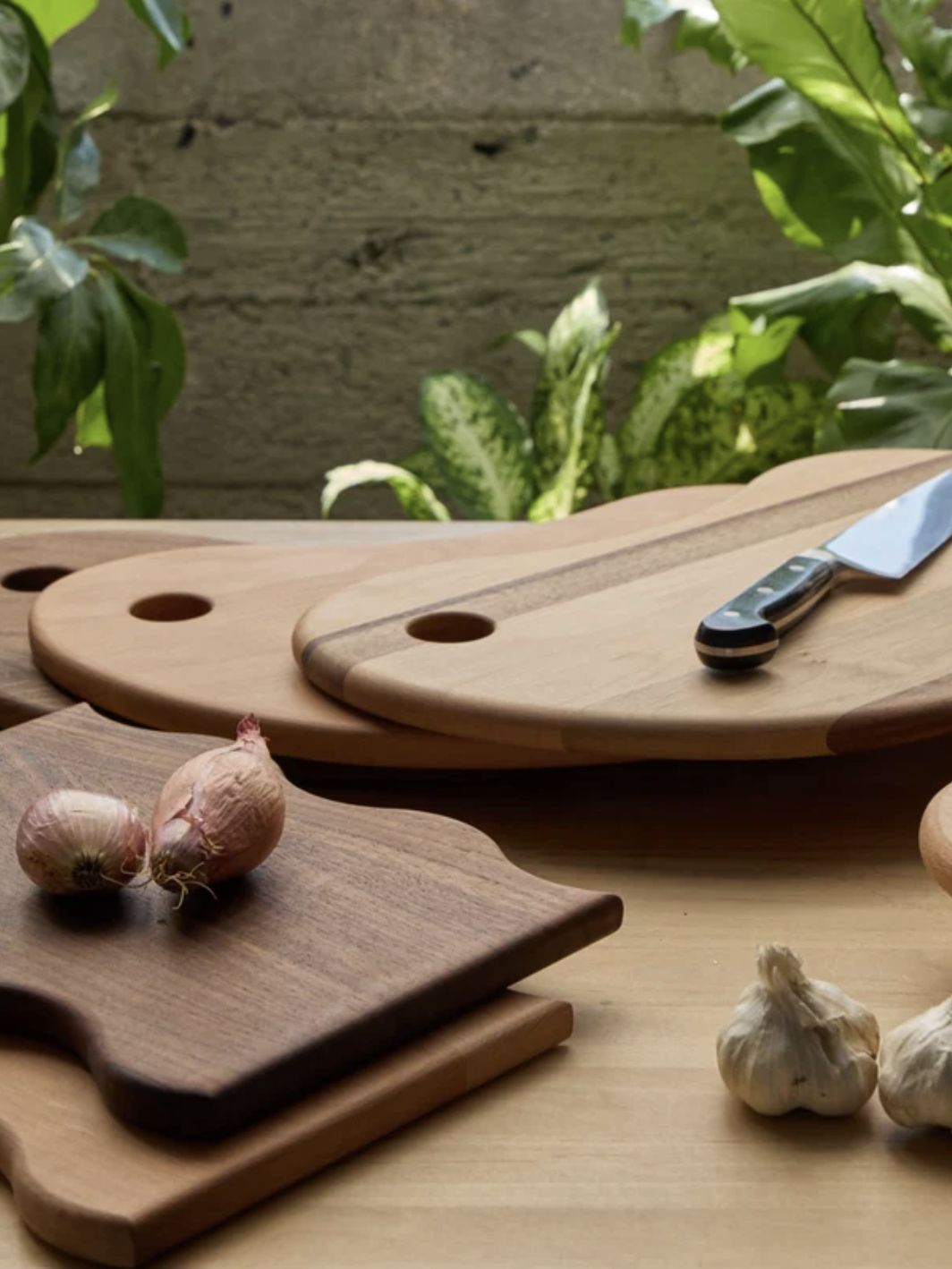 Wooden cutting boards and a knife on a table, surrounded by shallots, garlic, and artichokes, with green plants in the background.