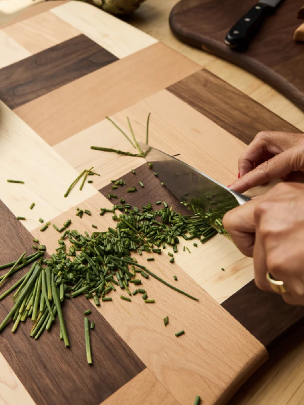 Hands chopping chives on a wooden cutting board with a knife. A bowl of cherry tomatoes is visible in the background.