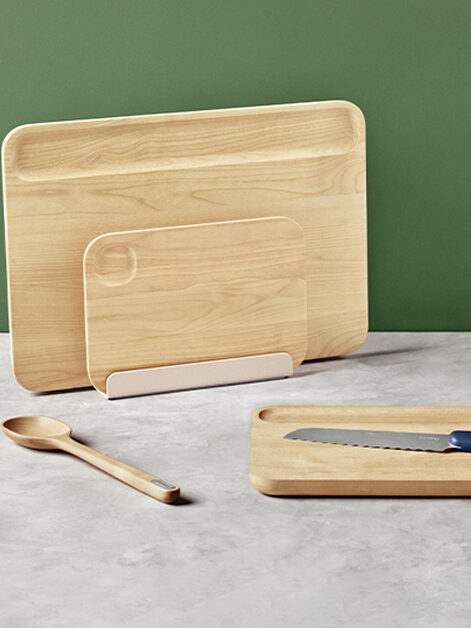 Wooden cutting boards, a tray with a serrated knife, and a wooden spoon on a gray countertop with a green background.