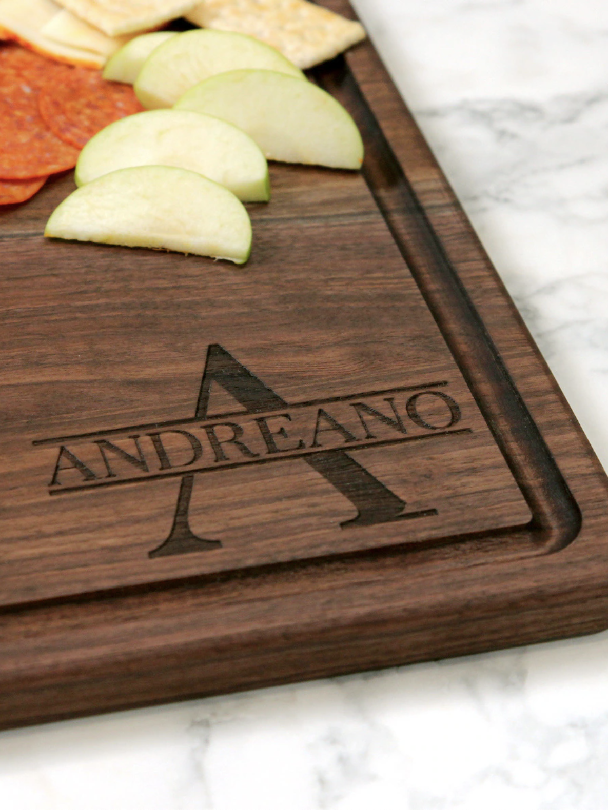 Wooden cutting board with the name "Andreano" and an engraved "A." Sliced apples, pepperoni, and cheese are on top.