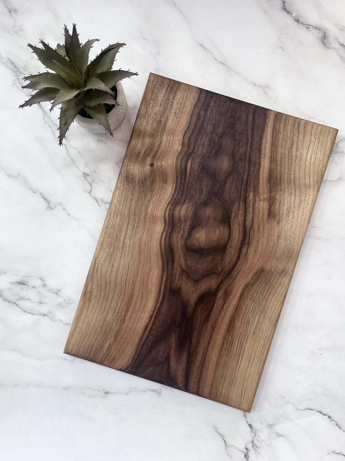 A wooden cutting board with a natural grain pattern is placed on a marble surface next to a small potted succulent.