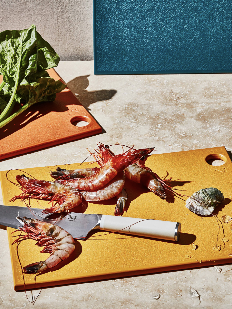 Prawns and a knife on an orange cutting board with a shell and leafy greens beside it; blue cutting board in the background.