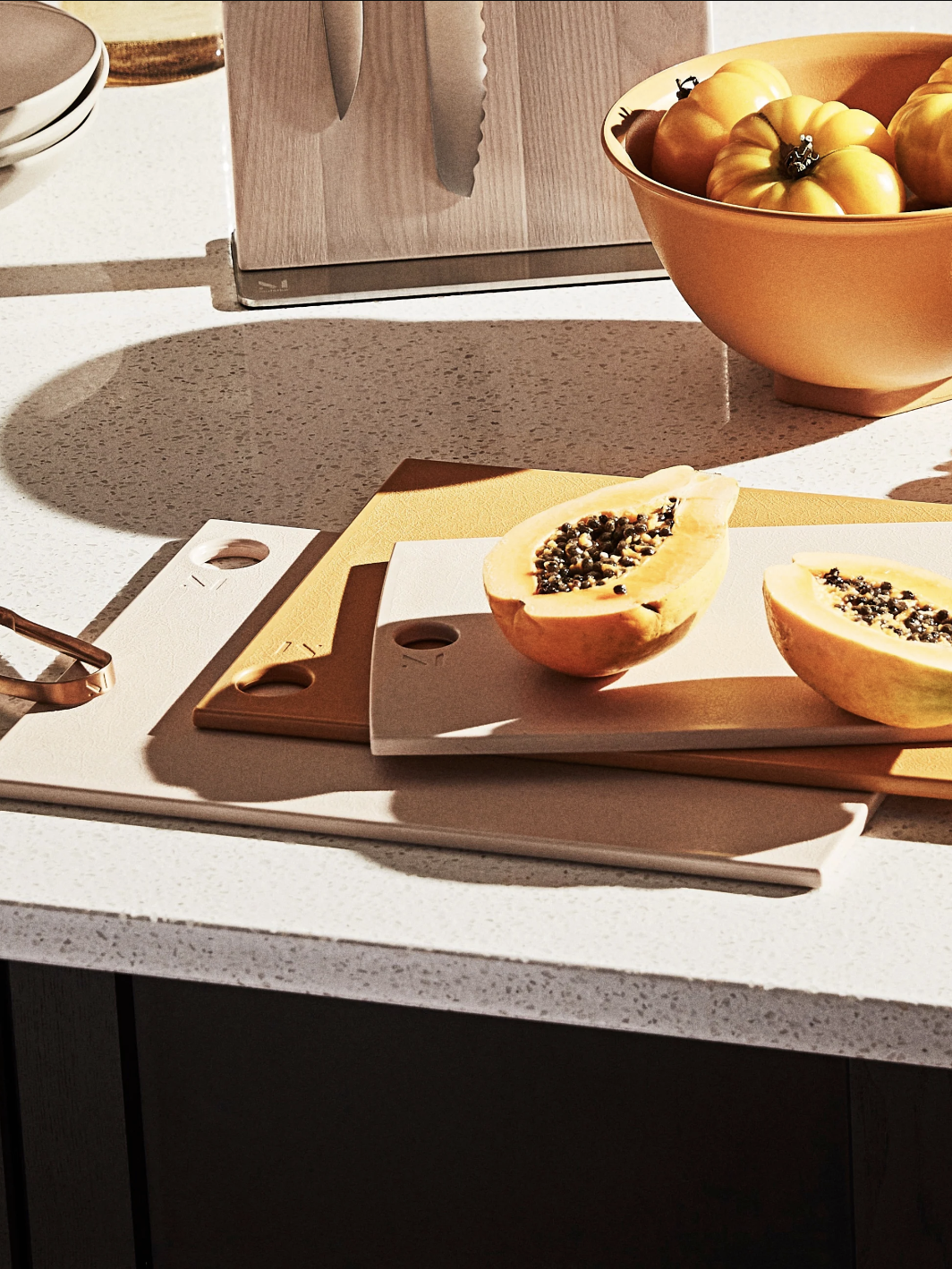 A halved papaya with seeds on a cutting board next to a knife and a bowl of yellow tomatoes on a kitchen counter.