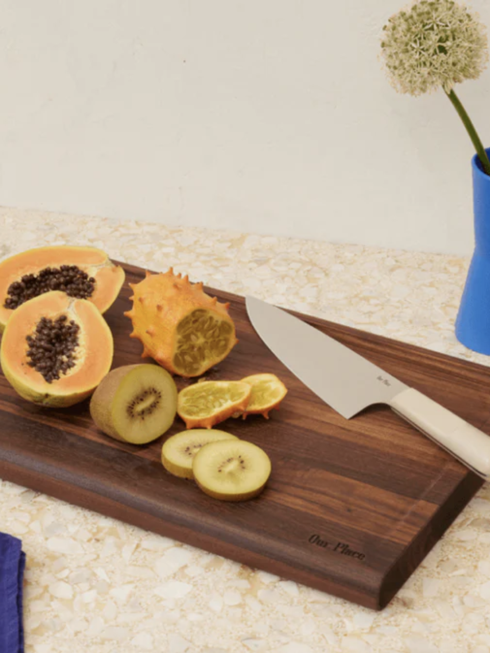 Fruits including papaya, kiwano, and sliced kiwi on a wooden cutting board with a knife, next to a blue vase with a flower on a speckled countertop.