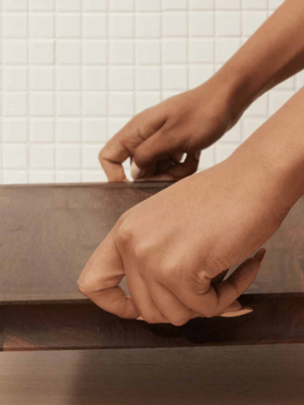 Hands adjusting a brown wooden box lid on a wooden surface with a white tiled background.