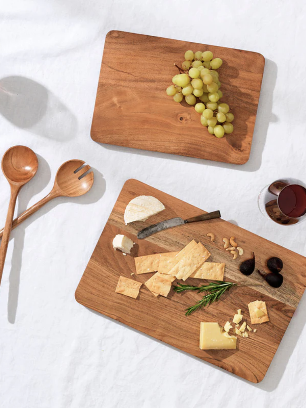 Cheese and fruit platter on wooden boards with grapes, cheese, figs, nuts, rosemary, utensils, and a glass of red wine on a white tablecloth.