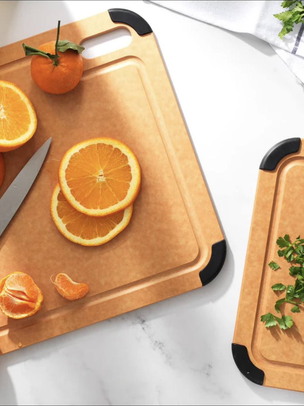 Two cutting boards on a white surface; one with sliced oranges and a knife, the other with chopped herbs. Adjacent whole orange and peeled segments.