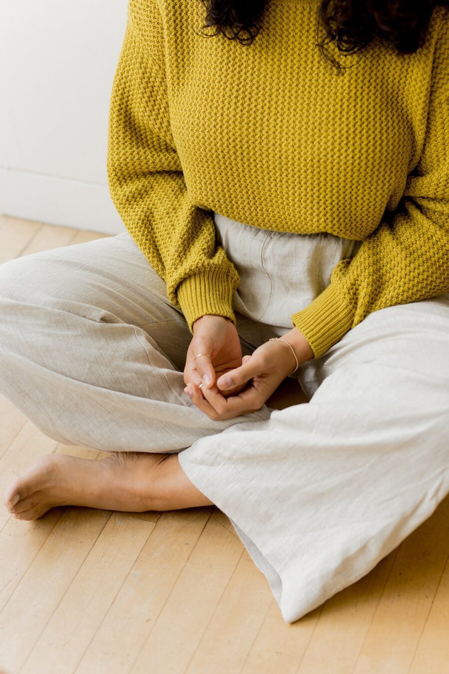 Person seated on the floor with crossed legs, wearing a textured yellow sweater and light trousers, hands resting in lap, barefoot on a wooden floor.