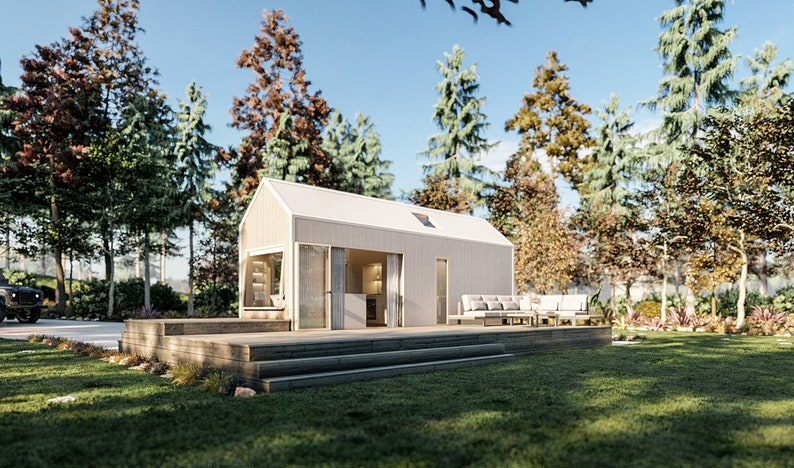 A modern tiny house with a wooden exterior stands on a raised deck in a forested area. The deck is furnished with outdoor seating. Trees surround the house, and the sky is clear and sunny.